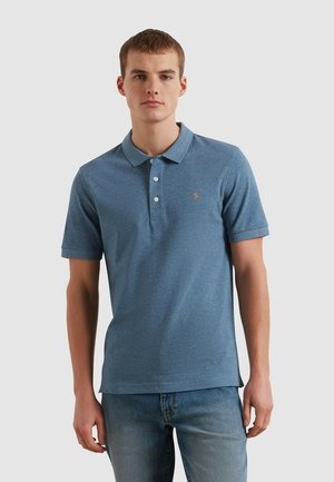 Junger Mann steht, trägt ein blaues kurzärmeliges Poloshirt mit Knöpfen und hellblaue Jeans vor einem einfarbigen Hintergrund.