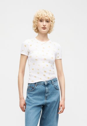 Donna bionda con capelli ricci che indossa una maglietta bianca a maniche corte con motivi di fiori gialli e jeans blu a vita alta.