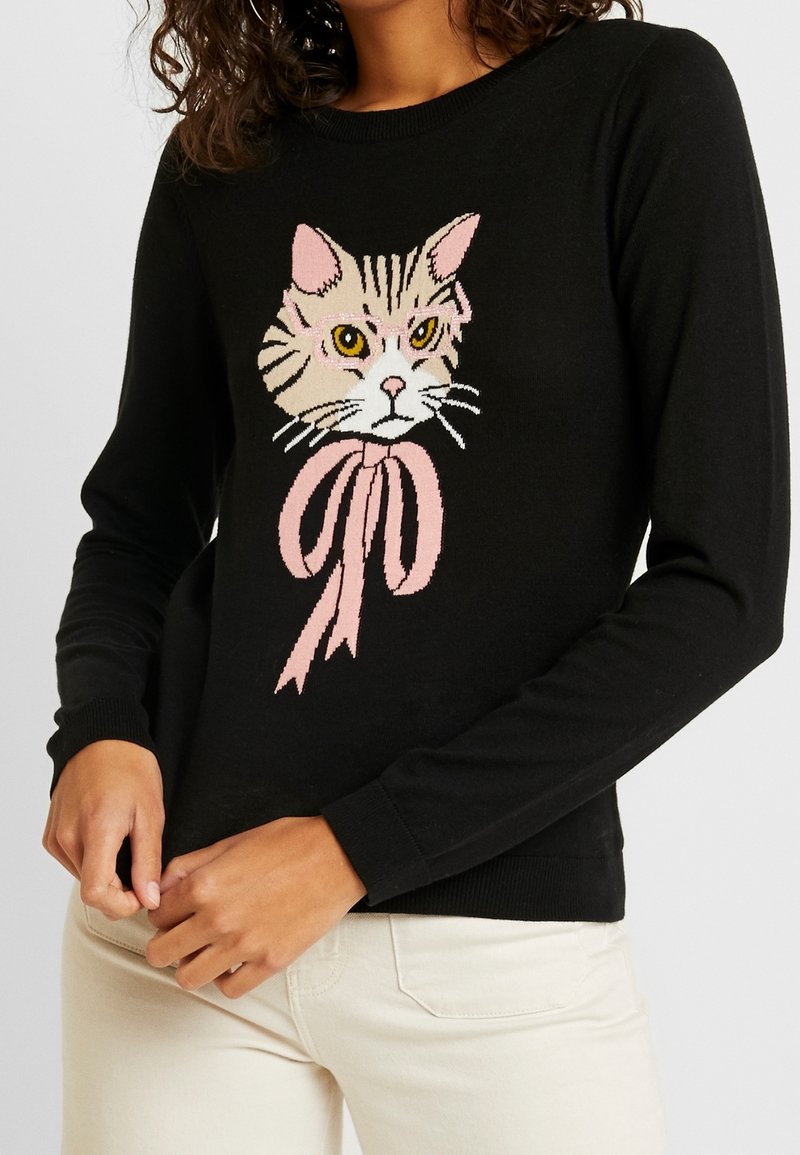 Pull noir à manches longues avec un visage de chat détaillé portant des lunettes roses et un grand nœud rose à l'avant, porté par une personne en pantalon clair.