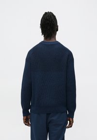 Tengerészkék kötött pulóver dombornyomott mintával, kerek nyakkivágással és bordázott mandzsettákkal. Hátulnézet, hozzáillő tengerészkék alsó résszel viselve.