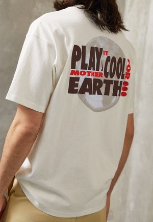 Henkilö, joka käyttää valkoista t-paitaa, jossa lukee "Play It Cool for Mother Earth" ja heikko maapallon kuva takana.