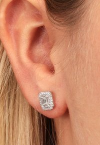 Silberner quadratischer Ohrstecker mit einem zentralen rechteckigen Stein, umgeben von kleinen klaren Steinen, vor einem hellen Hauthintergrund.