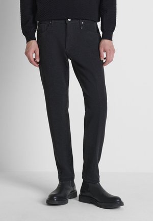 Man met strakke zwarte broek met voorzakken en zwarte leren Chelsea boots tegen een effen lichte achtergrond.
