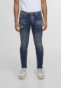 Blugi subțiri din denim albastru cu uzură la genunchi. Prezintă un design tradițional cu cinci buzunare și un sistem de închidere cu nasture.