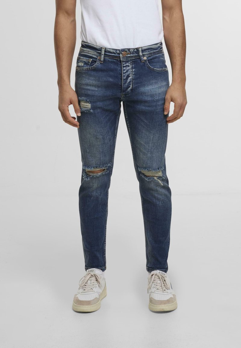 Blugi subțiri din denim albastru cu uzură la genunchi. Prezintă un design tradițional cu cinci buzunare și un sistem de închidere cu nasture.