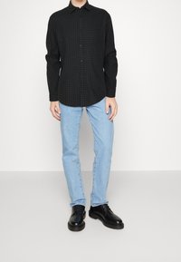 Camicia a quadri nera e verde, maniche lunghe, abbinata a jeans azzurri chiari e scarpe in pelle nere, in piedi contro uno sfondo neutro.