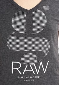 Šedé bavlněné tričko s výstřihem do V. Obsahuje velký bílý tečkovaný design loga a text "RAW" v tučném písmu, s menším textem níže.