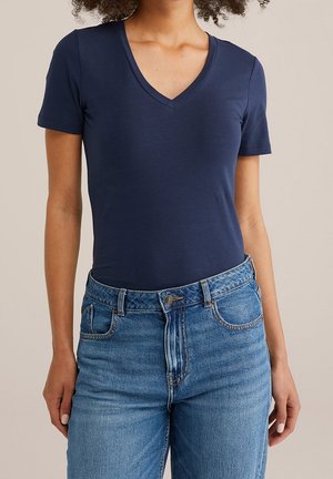 Osoba ubrana w granatowy shirt z krótkim rękawem i dekoltem w serek, włożony w wysokie niebieskie dżinsy, stojąca na tle bez wzoru.