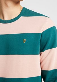 Sweter z okrągłym dekoltem w pasy, wyróżniający się mocnymi paskami w kolorze turkusowym i delikatnym różowym, z małym, haftowanym żółtym logo na piersi. Widoczna faktura dzianiny.