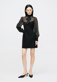 Μαύρο φόρεμα με διαφανή μακριά μανίκια και λαιμόκοψη με φιόγκο. Εφαρμοστή γραμμή με ίσια κοπή, απαλή υφή και χωρίς ορατά σχέδια.