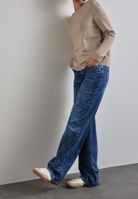 Person i beige tröja, blå vida jeans och vita sneakers som står mot en ljusgrå vägg med händerna i fickorna.