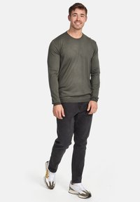 T-shirt à manches longues en olive, en tissu lisse, associé à un jean sombre et des baskets blanches avec des accents jaunes et gris.