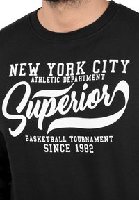 Czarna bluza z białym napisem. Wzór zawiera teksty: "NEW YORK CITY ATHLETIC DEPARTMENT", "Superior", "BASKETBALL TOURNAMENT" oraz "SINCE 1982".
