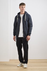 Navy zip-up hoodie with drawstring, paired with a white T-shirt and black pants, and white athletic shoes on wooden floor.