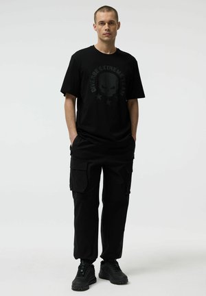 Mężczyzna stojący z rękami w kieszeniach, ubrany w czarną koszulkę z grafiką czaszki, czarne spodnie cargo oraz czarne sneakersy na jednolitym białym tle.