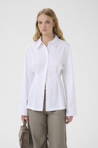 Young woman with long blonde hair wearing a white button-up shirt and taupe pants, holding a brown handbag in her left hand.