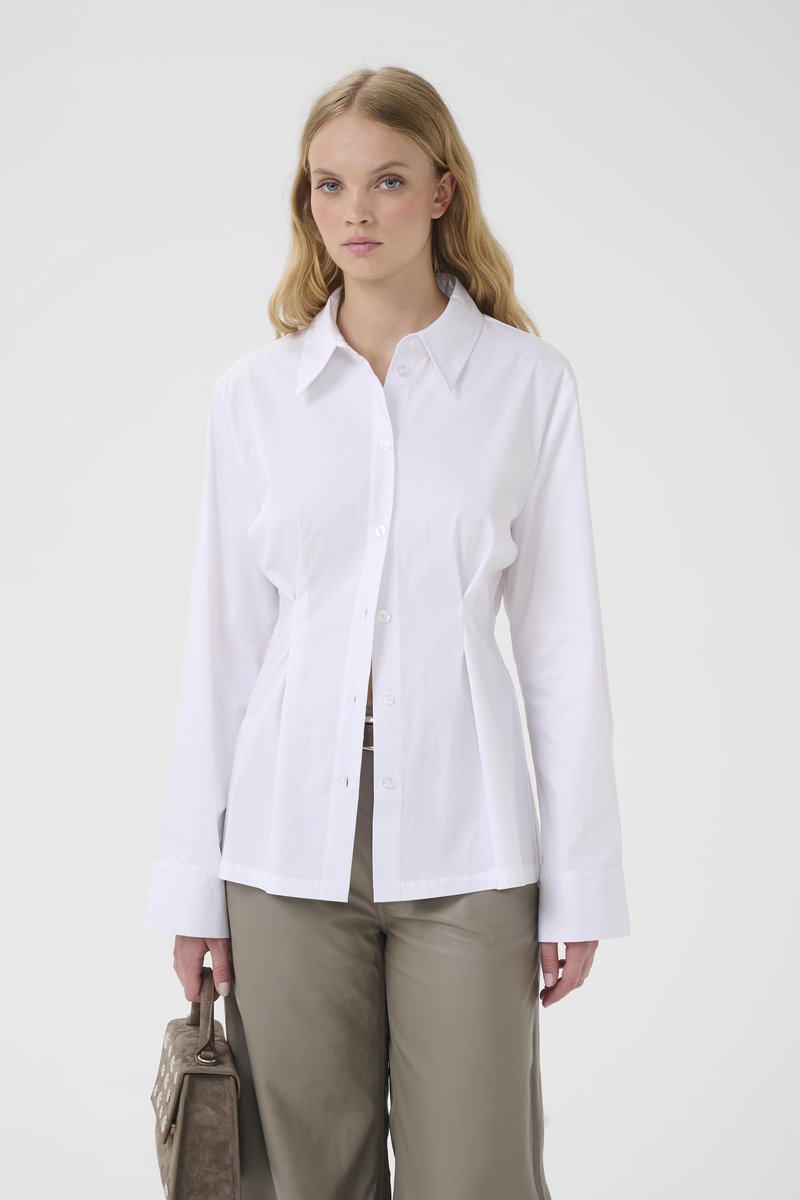 Young woman with long blonde hair wearing a white button-up shirt and taupe pants, holding a brown handbag in her left hand.