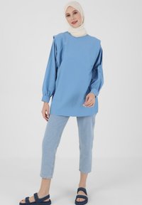 Maglietta oversized in cotone blu con maniche lunghe, abbinata a pantaloni cropped in denim chiaro e sandali in tessuto blu. Un hijab bianco avvolge la testa.