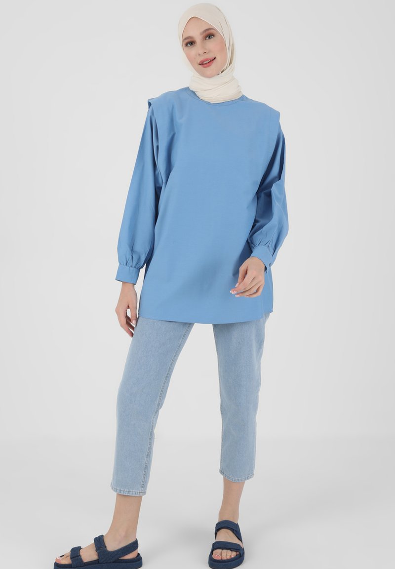 Maglietta oversized in cotone blu con maniche lunghe, abbinata a pantaloni cropped in denim chiaro e sandali in tessuto blu. Un hijab bianco avvolge la testa.