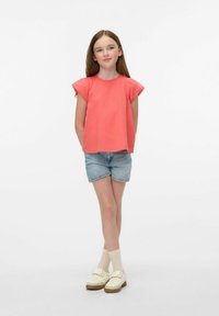 Top corto a maniche corte in corallo con tessuto testurizzato, abbinato a pantaloni corti in denim chiaro. Il modello indossa sneaker color crema e calze al ginocchio, in piedi con le braccia rilassate.