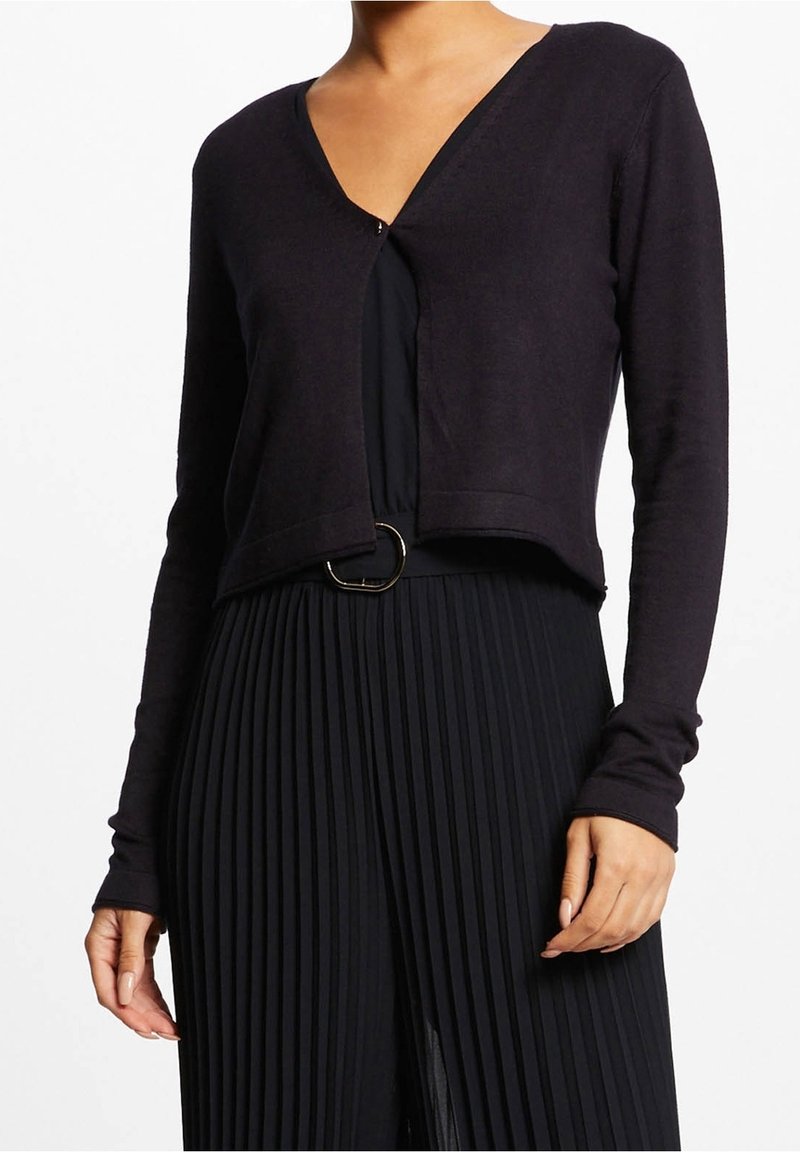 Femme portant un cardigan court noir par-dessus une robe plissée noire avec une ceinture à boucle ronde à la taille, visible du cou aux hanches.