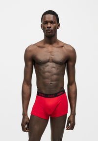 Αθλητικός άνδρας με σκουρόχρωμο δέρμα σε όρθια στάση, φοράει φωτεινά κόκκινα μποξεράκια με μαύρη ζώνη με την ένδειξη "HUGO", ουδέτερο γκρι φόντο.