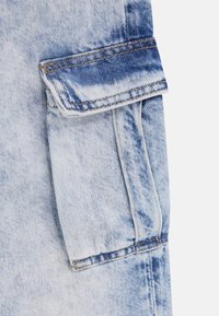 Svetlo modra denim tkanina ima žep s poklopcem. Material prikazuje zbledele, obrabljene teksture z nežnimi odtenki barve.