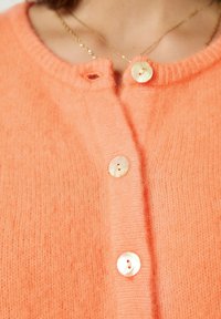 Persikinės oranžinės spalvos megztinis kardiganas su apvalia iškirpte, keturiais rausvais mygtukais ir minkštu paviršiumi. Nėra matomų raštų ar metalinių detalių.