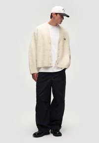 Cardigan in lana crema con finitura testurizzata, indossato sopra una maglietta bianca e abbinato a pantaloni cargo neri, completato da un cappellino bianco e nero.