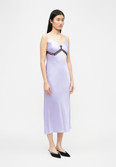 Γυναίκα που στέκεται φορώντας ένα λιλά σατέν φόρεμα με λεπτές τιράντες και μαύρη δαντελένια καρδιά, συνδυασμένο με μαύρα μυτερά φλατ παπούτσια.