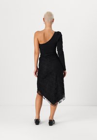 Μαύρο ολόσωμο κορμάκι με ένα μακρύ μανίκι και λεπτομέρεια ραχών, συνδυασμένο με μια ασύμμετρη φούστα από μαύρη δαντέλα που φτάνει μέχρι το μέσο της γάμπας. Μαύρα επίπεδα παπούτσια.