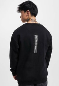 Black sweatshirt with a relaxed fit. Features a white text graphic on the back, ribbed cuffs, and a crew neckline. Soft fabric texture.