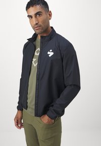 Svart lett jakke med glidelås, ståkrage og hvitt logo. Kombinert med en olivengrønn skjorte og matchende slim-fit bukser.