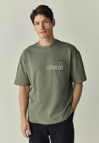 Olive spalvos marškinėliai trumpomis rankovėmis, tekstūruota medžiaga, apvalus kaklas ir priekinė kišenė. Ant krūtinės spausdinamas žodis "LESOLEIL" šviesia spalva.