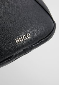 Borsa in pelle nera con superficie testurizzata, caratterizzata da un logo "HUGO" argentato e un sottile design curvo lungo il bordo.