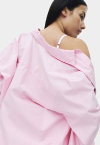 Chemise oversize rose clair en tissu lisse, dotée d'un col classique et de rayures verticales subtiles, portée partiellement sur l'épaule.