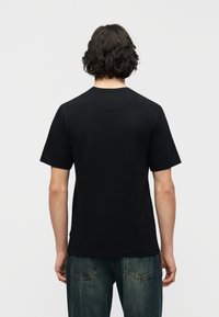 Άτομο με σκούρα σγουρά μαλλιά μέ χρι τους ώμους, που φορά ένα απλό μαύρο κοντομάνικο μπλουζάκι και μπλε τζιν, με την πλάτη γυρισμένη.