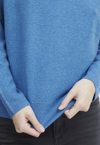 Pull en tricot bleu avec des poignets et un ourlet côtelés, présentant une texture subtile. La personne ajuste l'ourlet, mettant en valeur son ajustement.