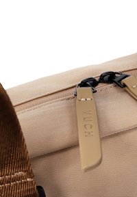 Beige Tasche aus Stoff mit glatter Oberfläche, ausgestattet mit schwarzen Reißverschlüssen und hellbraunen Reißverschlussanhängern, die mit "YUCH" graviert sind. Ein brauner Träger ist auf dem Bild sichtbar.