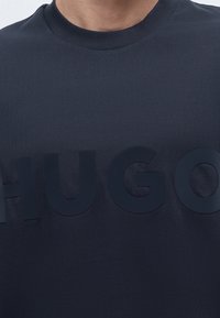 Άτομο που φοράει σκούρο μπλε πουκάμισο με μεγάλα ανάγλυφα γράμματα που σχηματίζουν το "HUGO" στο στήθος.