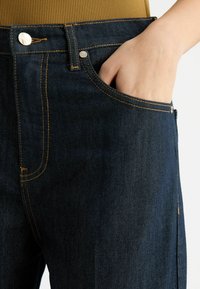 Jeans di denim blu scuro con cuciture arancioni, design a cinque tasche e chiusura con bottone metallico. Presenta un primo piano di una mano nella tasca anteriore.