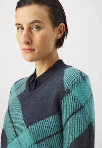 Ullgenser med mørk marineblå bunn, med turkise diamantmønstre og svarte skrå striper, designet med V-hals og krage.