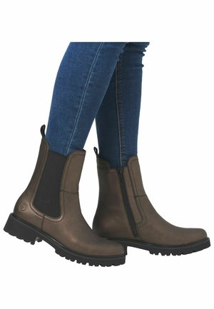 Botas de cuero marrón hasta el tobillo con paneles elásticos en los laterales, una cremallera y suelas de goma negra texturizada, combinadas con jeans de mezclilla azules.