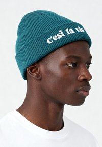 Cappellino in maglia color verde acqua con texture a coste e bordo piegato, con la scritta "c’est la vie" ricamata in bianco. Il modello indossa una maglietta bianca.