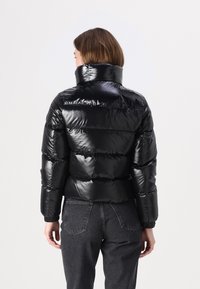 Zwarte pufferjack met een glanzende, gewatteerde afwerking. Hoge kraag en aansluitende mouwen. Gekleed met donkere spijkerbroek. Achteraanzicht weergegeven.