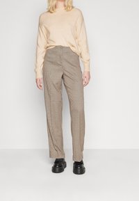 Pull beige en tricot associé à un pantalon taille haute à carreaux marron et blanc. Des chaussures noires épaisses complètent la tenue.