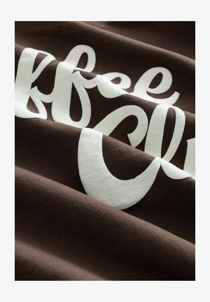 Bruine katoenen stof met witte, gestructureerde letters. Het ontwerp bevat vloeiende letters in een sierlijk schrift en vetgedrukte lettervormen, wat de zachtheid van de stof benadrukt.