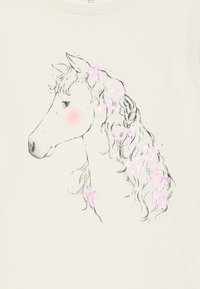 Hvitt t-skjorte med et svart og rosa glitrende grafikk av et hodeskjelett av en hest med en flettet manke pyntet med rosa sløyfer.