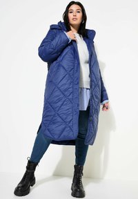 Abrigo largo y acolchado en azul marino con acolchado en forma de diamante, con capucha y cremallera delantera, llevado sobre un suéter gris y jeans azules. Botas negras gruesas.