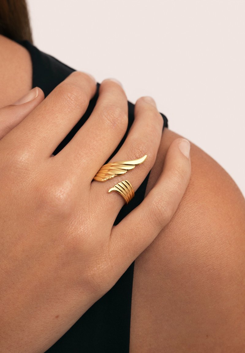 Anillo de color oro con un diseño de alas y surcos; llevado en una mano de tono de piel medio sobre un fondo negro.